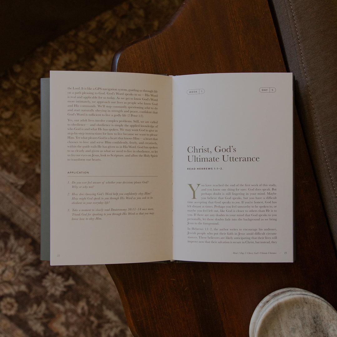 An open book laid on a dark brown leather surface, showing printed text pages with the title 'Christ, God's Ultimate Utterance' on the right page. A small round metal container is partially visible at the lower right.
