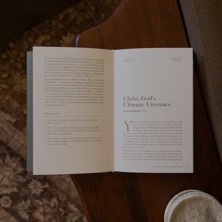 An open book laid on a dark brown leather surface, showing printed text pages with the title 'Christ, God's Ultimate Utterance' on the right page. A small round metal container is partially visible at the lower right.