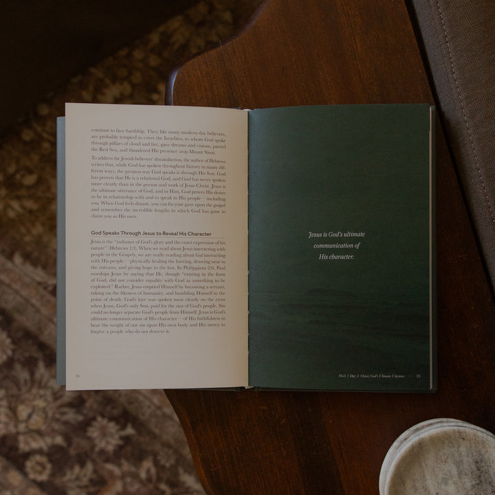 An open book spread on a wooden table, with printed text on both pages and a dark green title page visible. A cup rim and fabric surface are partially visible around it.