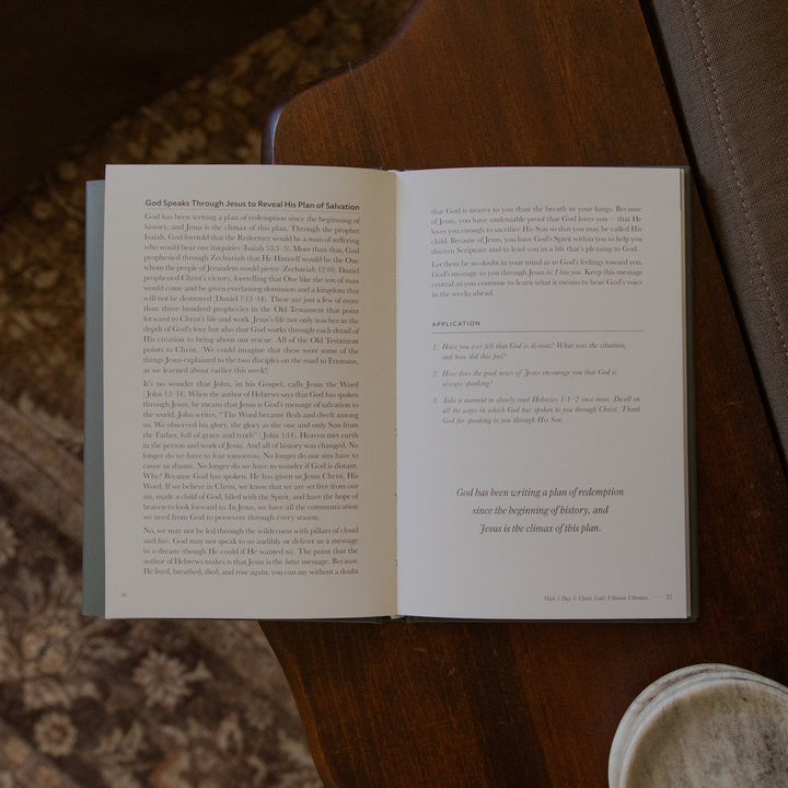 An open Bible book showing two printed pages with text, photographed from above on a brown textured surface next to a white cup.