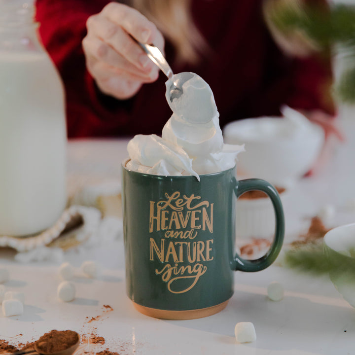 Christian woman putting whipped cream in Christmas mug that says Let Heaven and Nature Sing