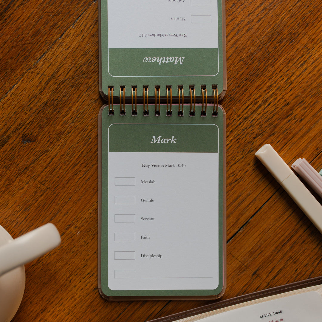 Highlighting through Scripture flipbook flipped to the Mark Card near an open bible on a wooden table near some highlighters