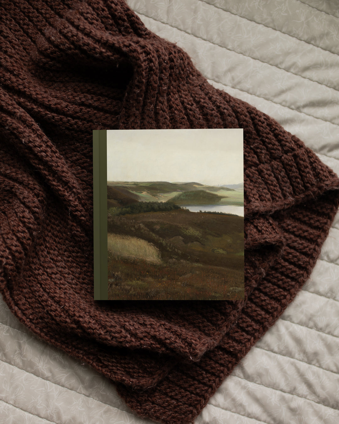 Brown knitted blanket draped over a textured surface with a small landscape painting on top.