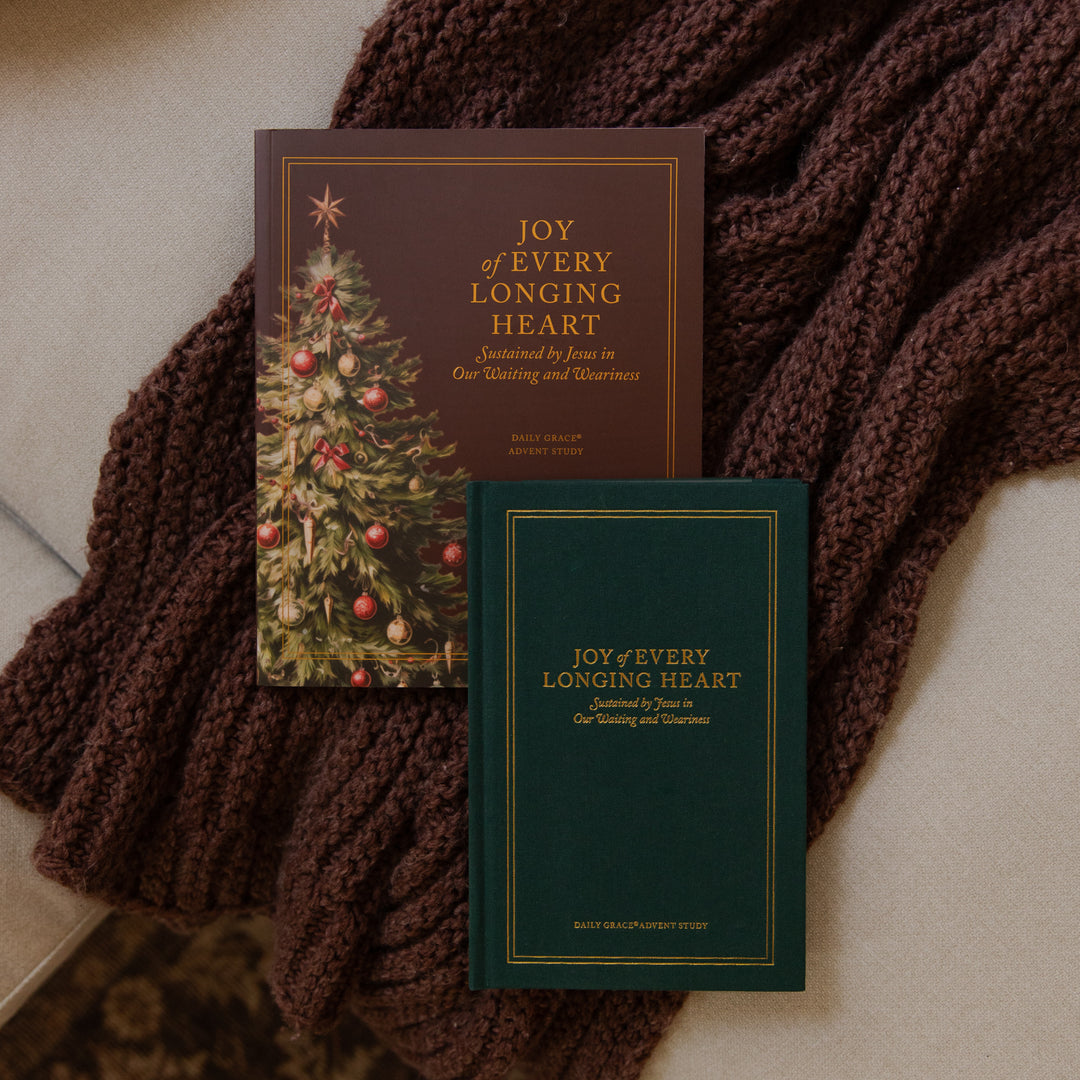 Two copies of the book 'Joy of Every Longing Heart' arranged on a beige surface with a chunky brown knit blanket. One copy has a dark brown cover with a Christmas tree illustration, and the other has a dark green cover with gold lettering.