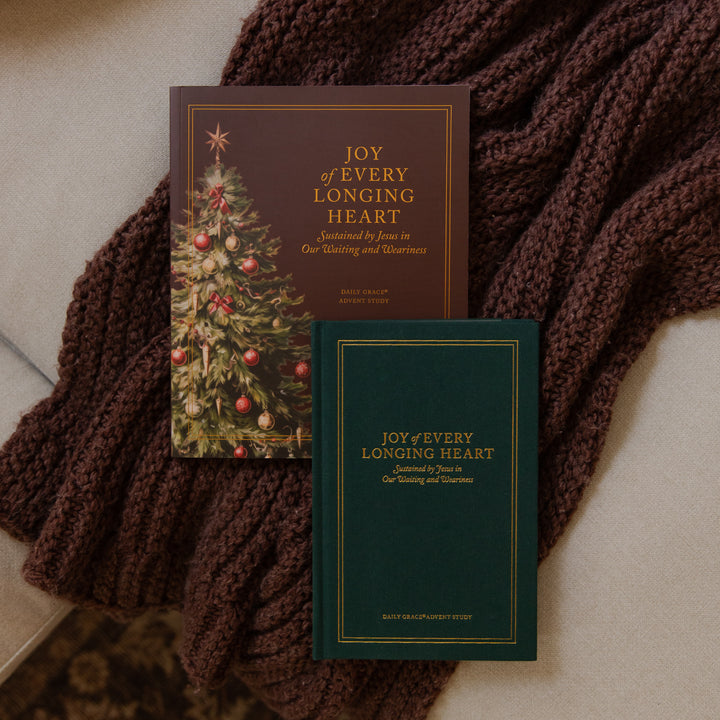 Two copies of the book 'Joy of Every Longing Heart' arranged on a beige surface with a chunky brown knit blanket. One copy has a dark brown cover with a Christmas tree illustration, and the other has a dark green cover with gold lettering.