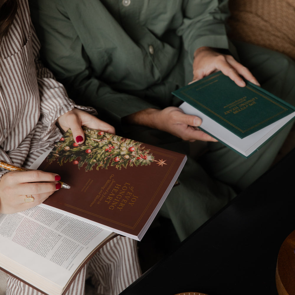 Two people seated indoors holding and reading decorative books. One person in striped pajamas holds a brown book with a Christmas tree illustration, while another person in an olive green outfit holds a dark green hardcover book.