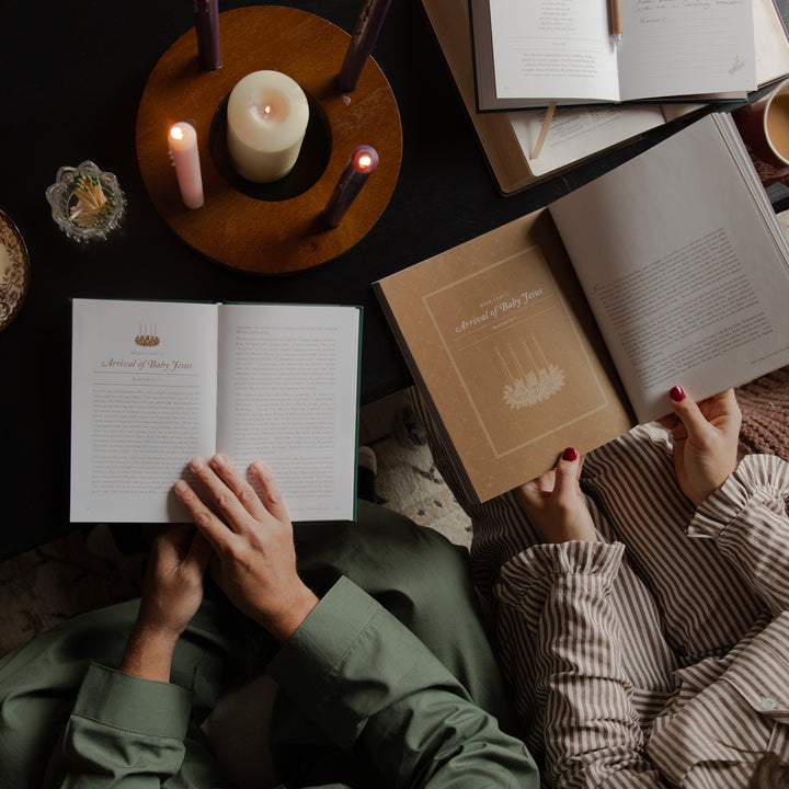 An overhead lifestyle shot of two people sitting at a dark table, reading cozy books with lit candles. One person wears an olive green shirt and holds an open book, while the other wears striped pajamas and holds a brown kraft paper book.