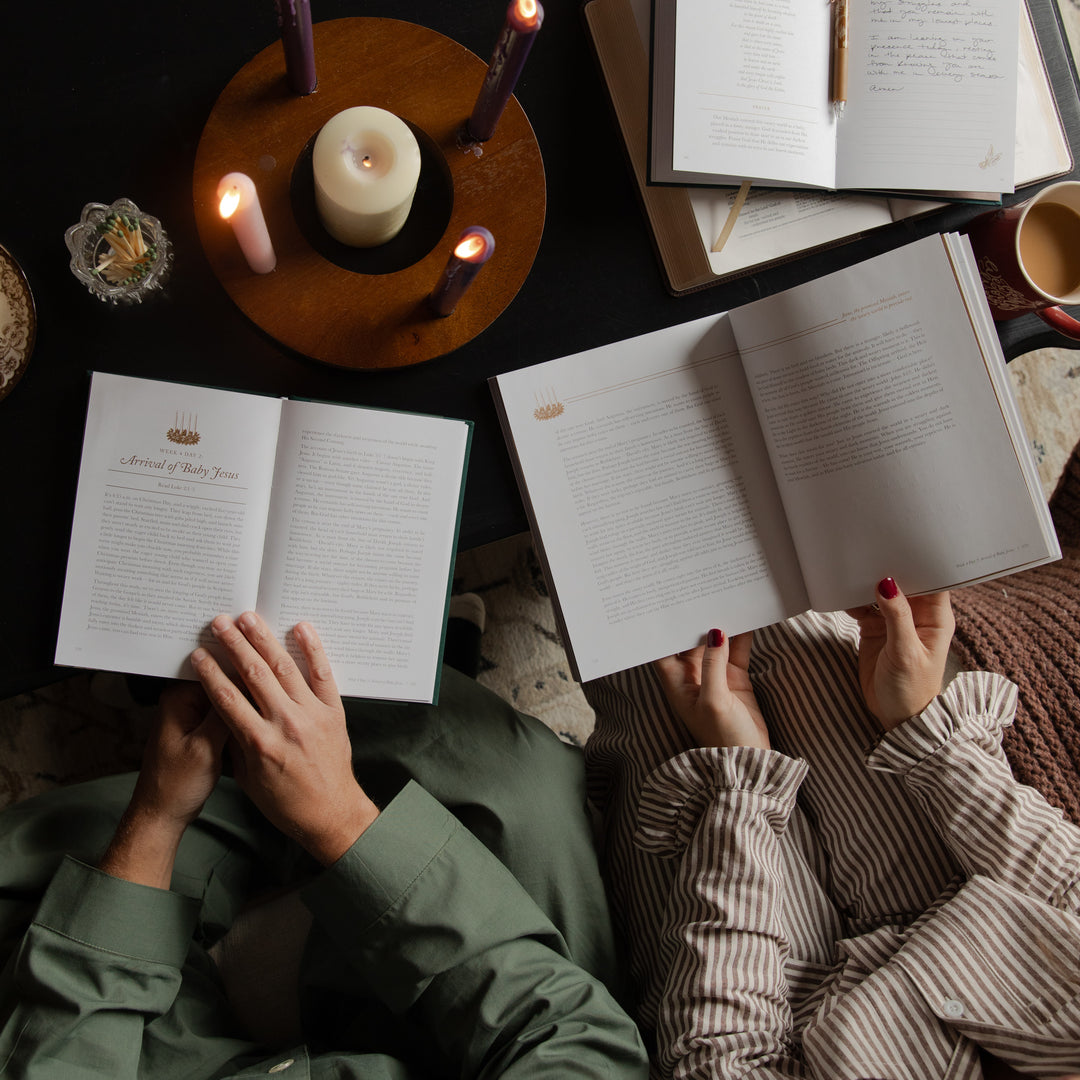 Overhead view of two people reading open books on a dark table, surrounded by lit candles, with one person wearing an olive green shirt and the other wearing a beige and brown striped top.
