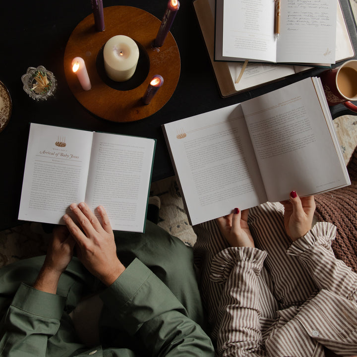 Overhead view of two people reading open books on a dark table, surrounded by lit candles, with one person wearing an olive green shirt and the other wearing a beige and brown striped top.
