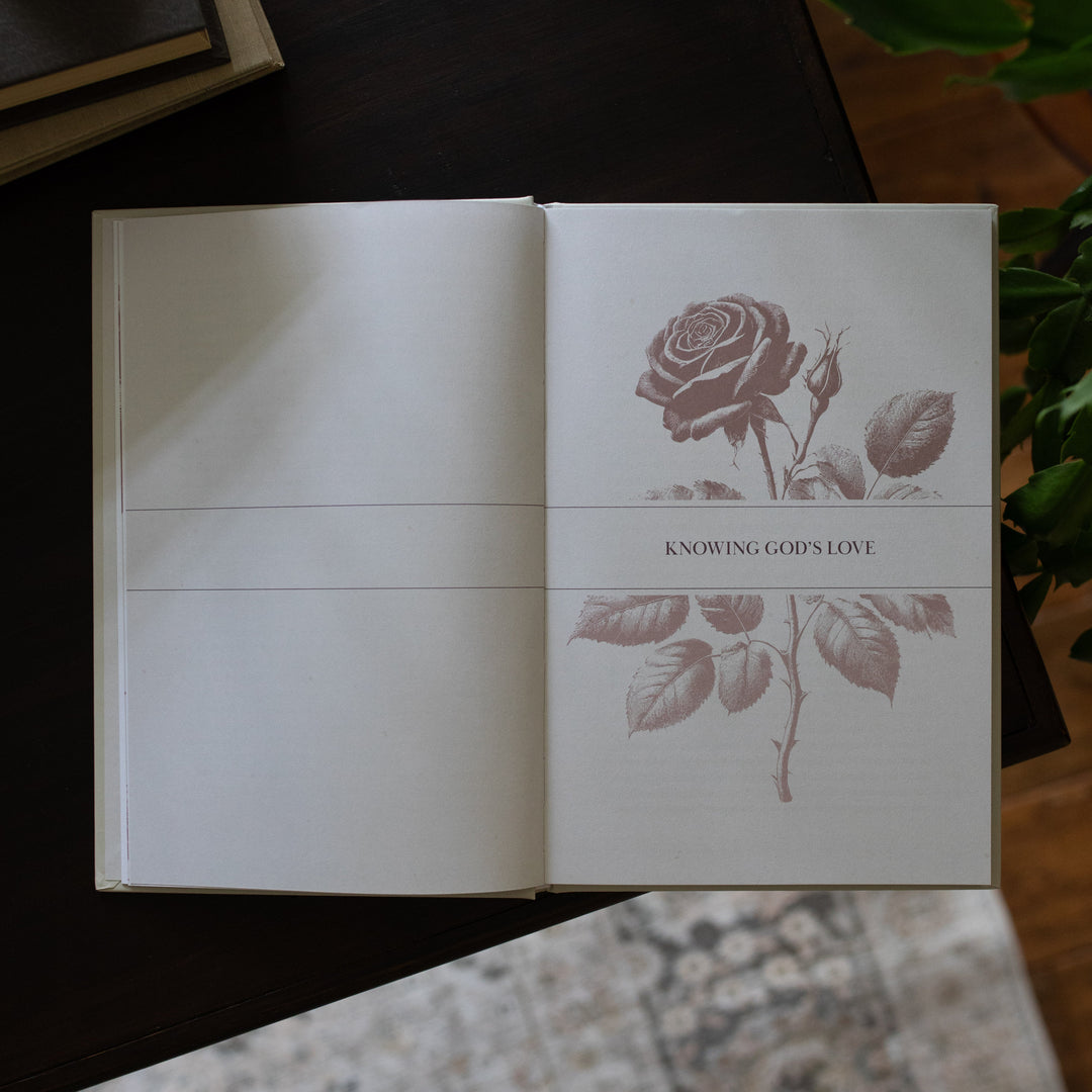His Love Endures Forever book opened to the \Knowing God's Love page on a dark wooden table with some greenery nearby