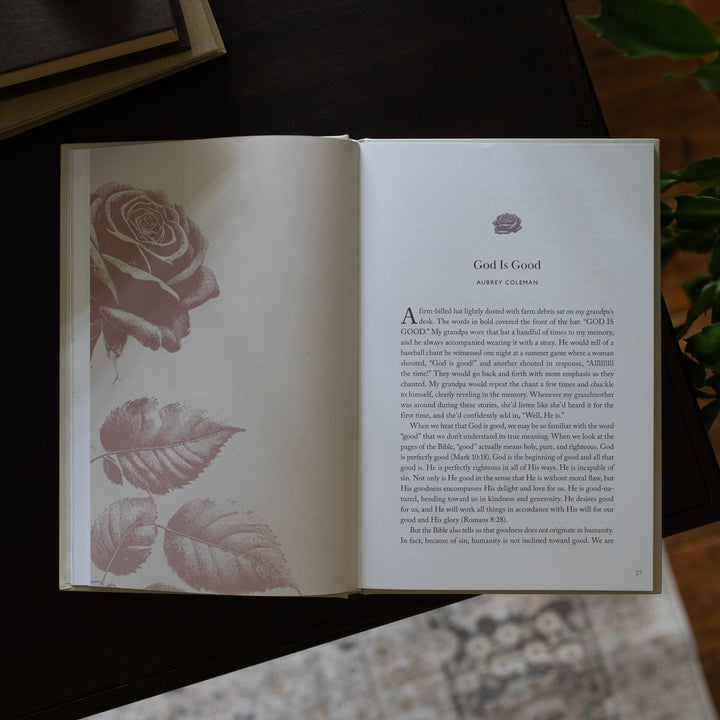 His Love Endures Forever book opened to the God is Good page on a dark wooden table with some greenery nearby