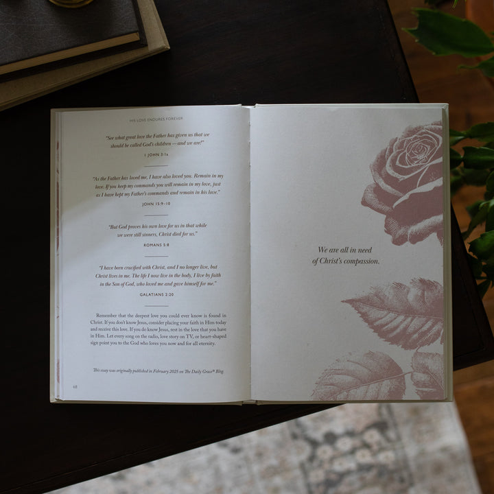 His Love Endures Forever book opened to the We are all in need of Christ's compassion page on a dark wooden table with some greenery nearby