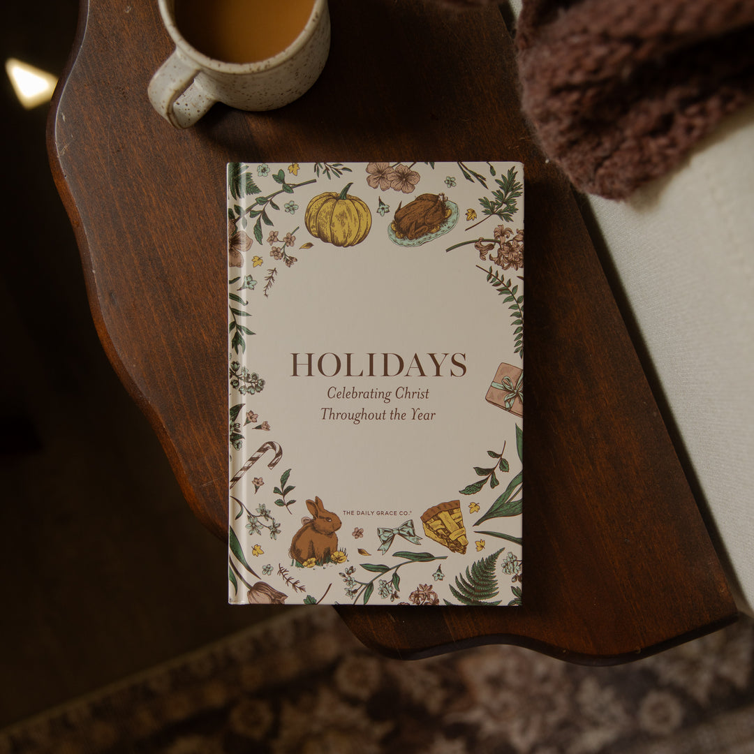 A hardcover book titled 'Holidays: Celebrating Christ Throughout the Year' by Dr. Bill Swart, styled on a dark wooden table next to a ceramic mug and a cozy brown knit blanket.