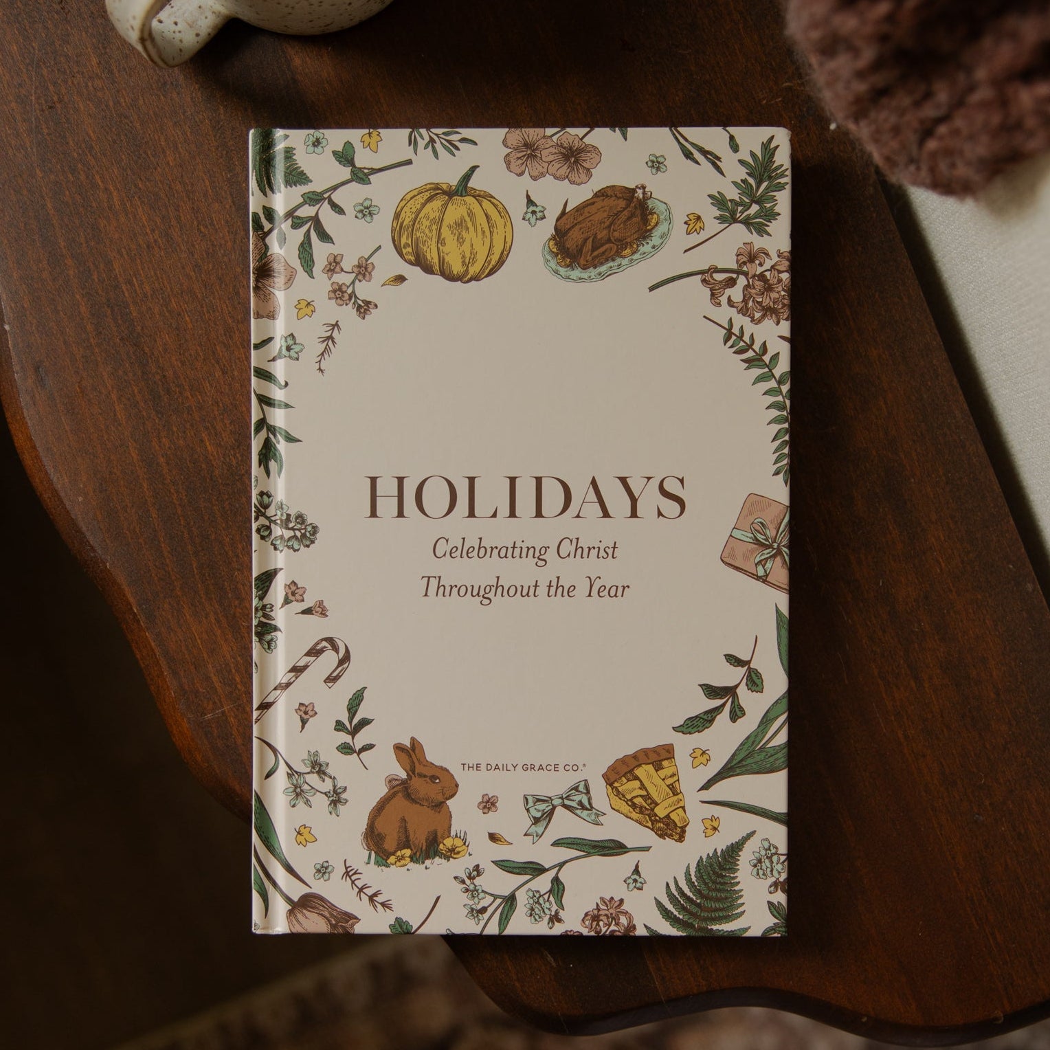 A hardcover book titled 'Holidays: Celebrating Christ Throughout the Year' by Dr. Bill Swart, styled on a dark wooden table next to a ceramic mug and a cozy brown knit blanket.