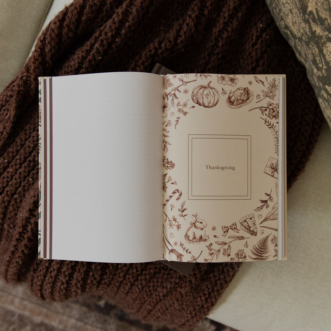 An open book or journal laid on a brown knitted blanket, showing a blank white left page and a decorated right page with autumn illustrations and the word 'Thanksgiving' in a central box.
