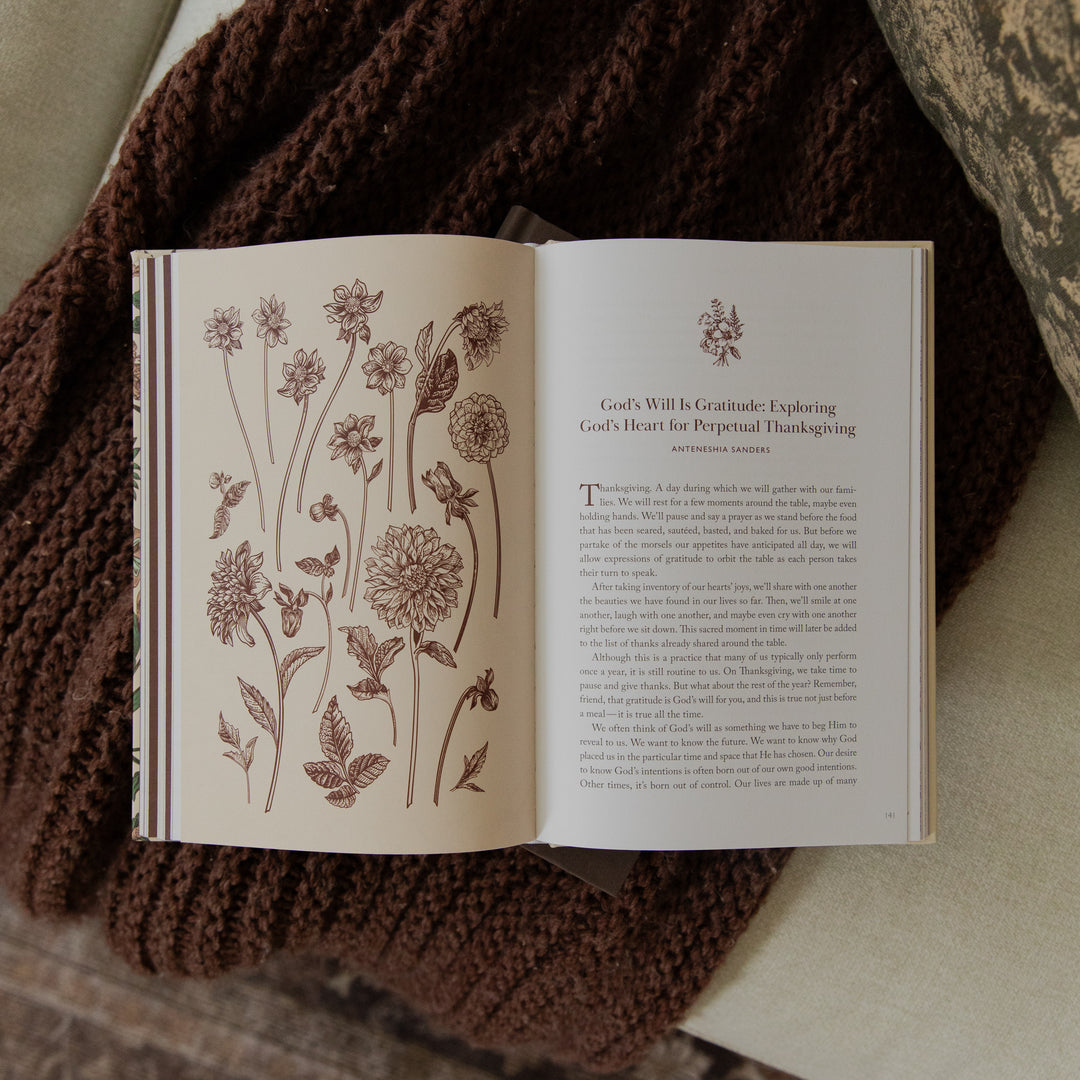 An open book with illustrated floral pages laid on top of a brown knitted blanket, photographed from above on a neutral fabric surface.