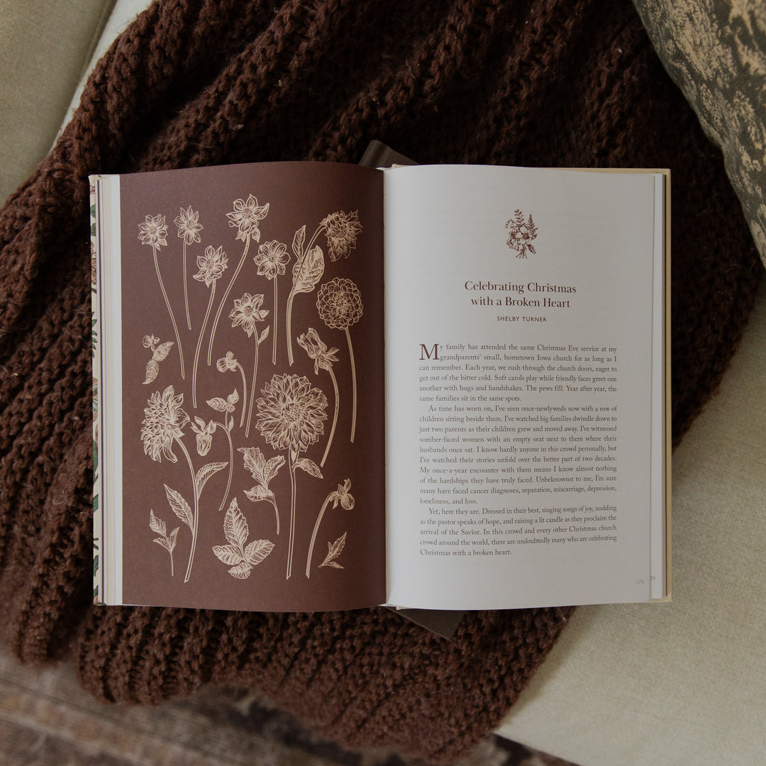 An open book laid on a brown knit blanket, showing a floral illustrated left page and a text page with the heading 'Celebrating Christmas with a Bordered Heart' on the right page.