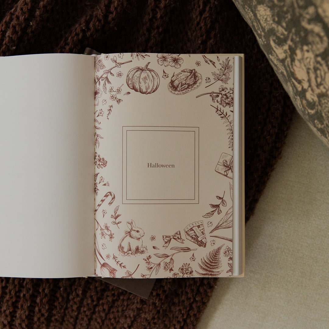An open book or journal showing a cream-colored inner page with delicate brown Halloween-themed illustrations, including pumpkins, bats, and autumn foliage, with the word 'Halloween' centered inside a double-line border. The book rests on a dark brown knitted blanket.