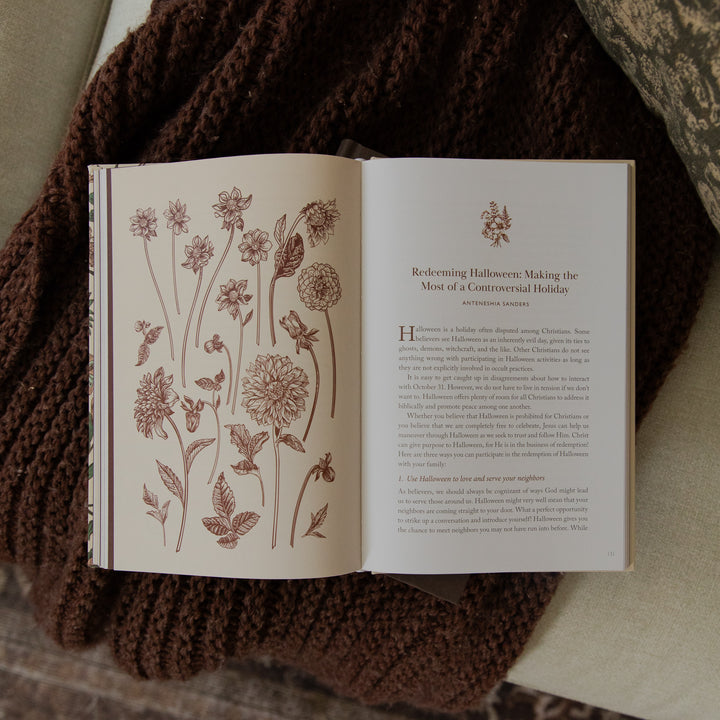 An open book with botanical illustrations and text on the pages, placed on top of a dark brown knitted blanket.