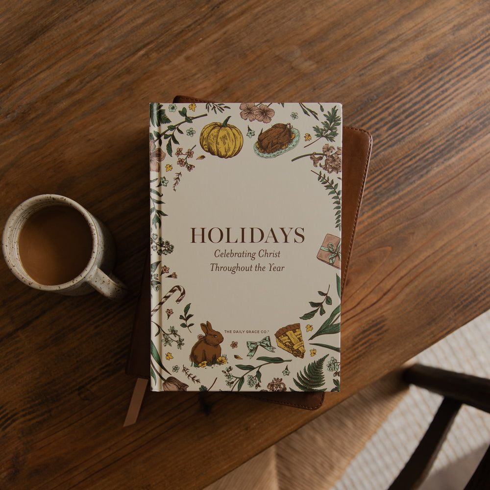 A hardcover journal titled 'HOLIDAYS: Celebrating Christ Throughout the Year' by The Daily Grace Co, featuring an autumn-themed floral border illustration. The journal is displayed on a wooden table next to a ceramic coffee mug.