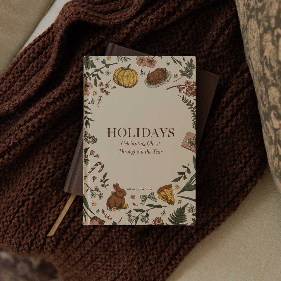 A holiday-themed book or journal titled 'HOLIDAYS Celebrating Christ Throughout the Year' by Holy Hands Press, styled on top of a dark brown chunky knit blanket.