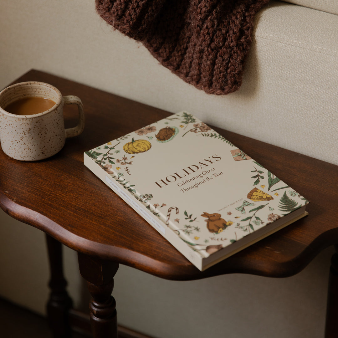 A hardcover book titled 'HOLIDAYS: Creating Cards Throughout the Year' resting on a dark wooden side table next to a ceramic mug of coffee, with a cozy brown knit blanket draped over a light beige sofa in the background.