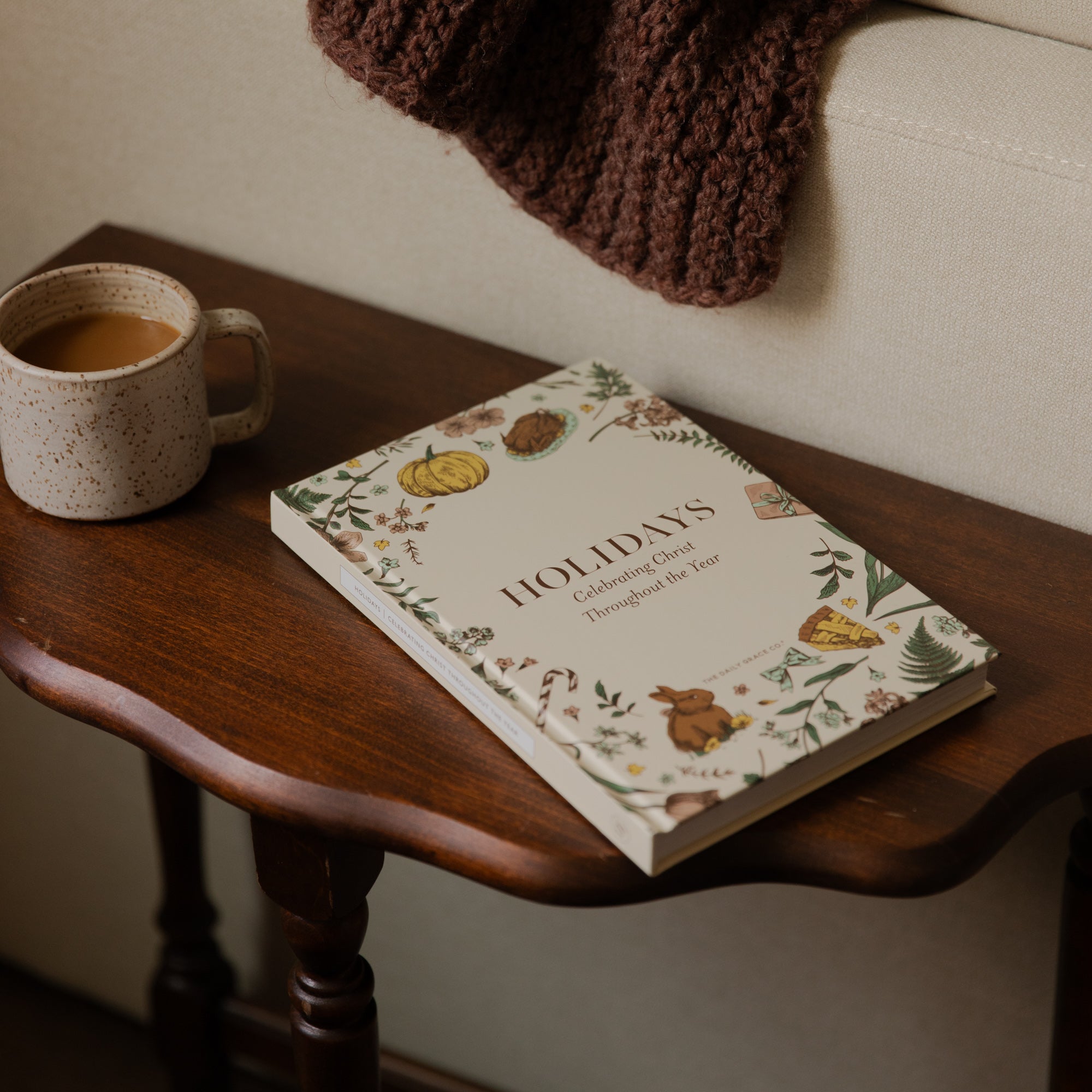 A hardcover book titled 'HOLIDAYS: Creating Cards Throughout the Year' resting on a dark wooden side table next to a ceramic mug of coffee, with a cozy brown knit blanket draped over a light beige sofa in the background.