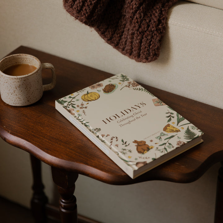 A hardcover book titled 'HOLIDAYS: Creating Cards Throughout the Year' resting on a dark wooden side table next to a ceramic mug of coffee, with a cozy brown knit blanket draped over a light beige sofa in the background.