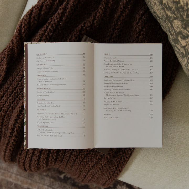 An open book showing a printed table of contents, photographed from above on a soft beige fabric surface with a brown knit blanket nearby.