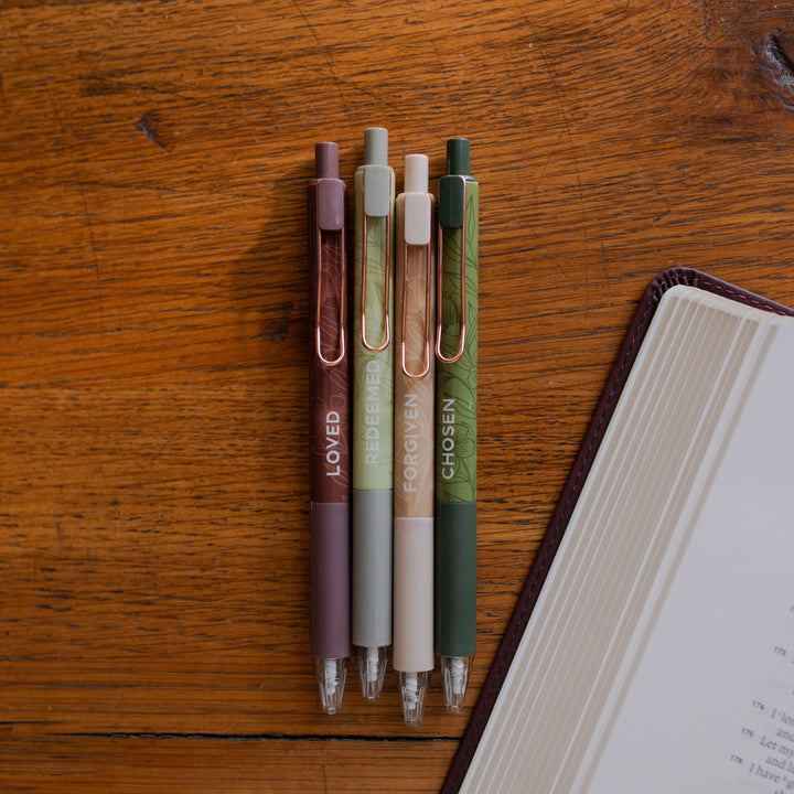 All four of the Identity Pen Set on a wooden table next to a corner of an open bible