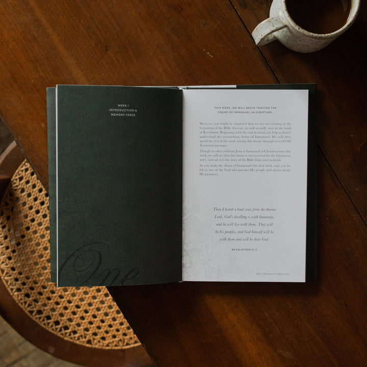 The Immanuel Hardcover opened to the Week 1 introduction page on a wooden table next to a coffee cup