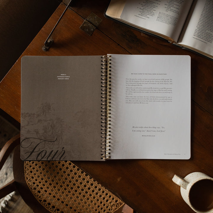 The Immanuel Spiral cover opened to the week 4 introduction and memory verse page on a wooden table near an open bible and a coffee cup