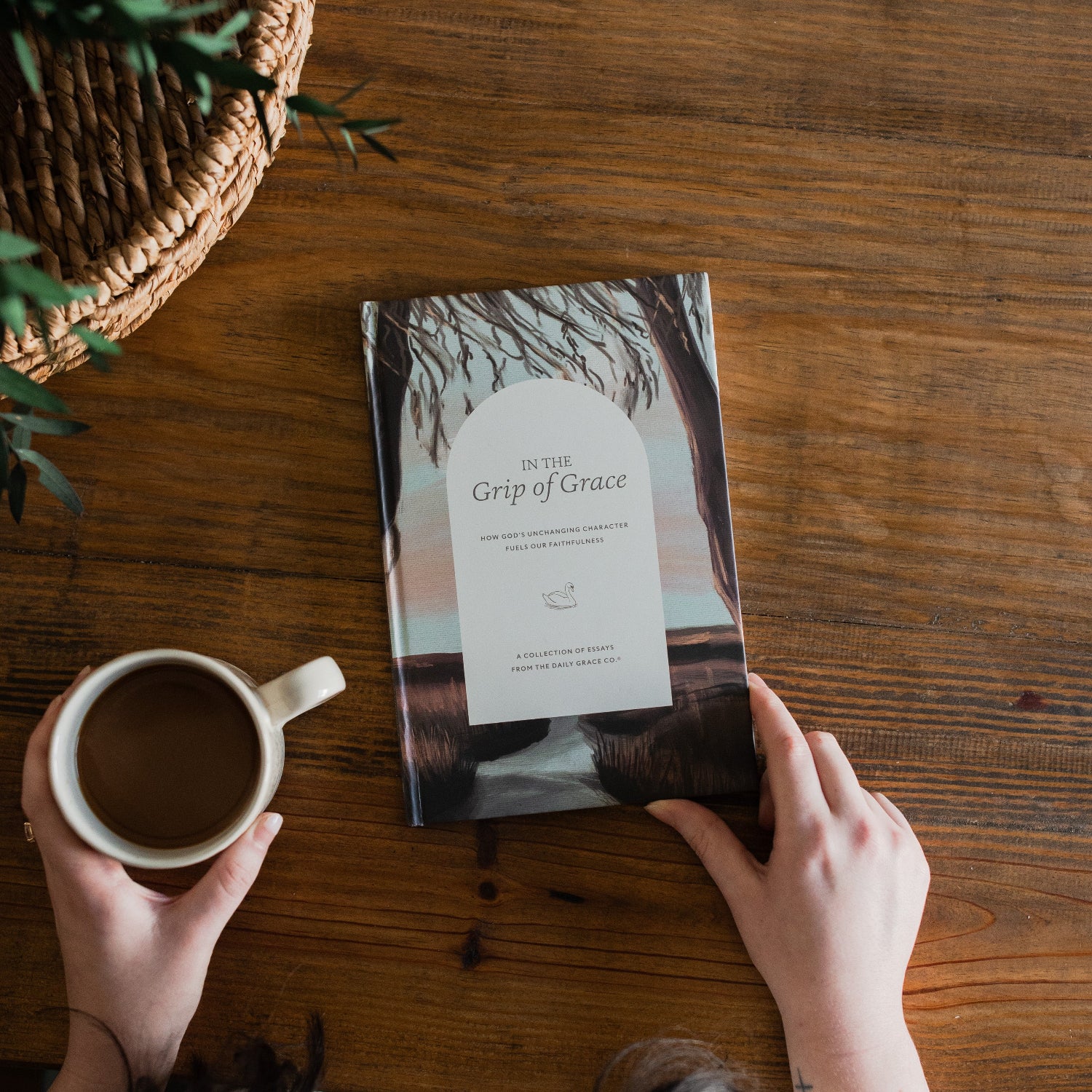 In the Grips of His Grace Study cover on a brown wooden table next to a woven basket and a white coffee cup with woman's hands holding the cup and the book on the corner