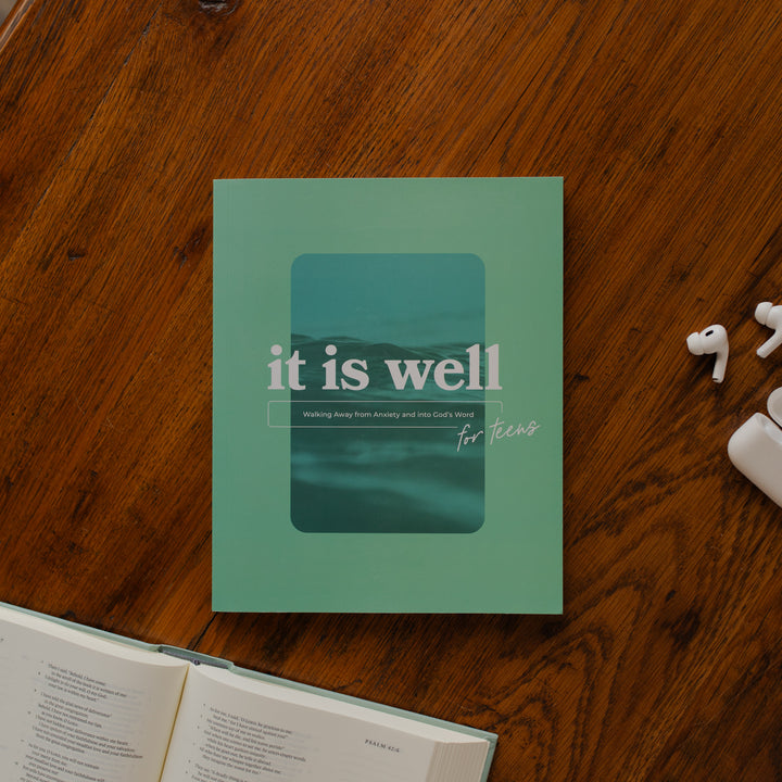 It is Well Teen Study cover on a wooden table next to an open bible and some apple earbuds