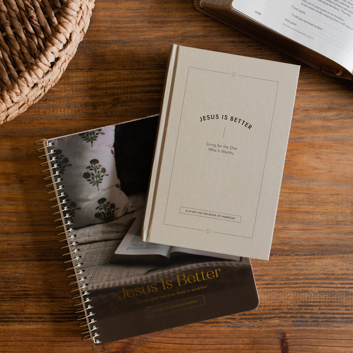 Jesus is Better Men and Women's Study cover tilted to an angle on a wooden desk next to a woven basket and an open bible