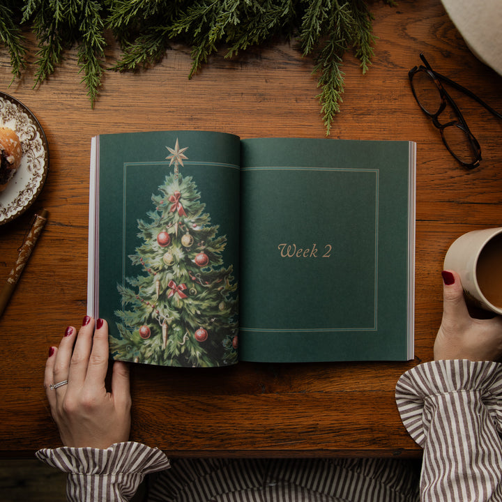 An open journal or book on a wooden table, showing a decorated Christmas tree illustration on the left page and the words 'Week 2' on the right page. A person's hands with red nail polish hold the open book, with a mug, glasses, and evergreen branches around it.