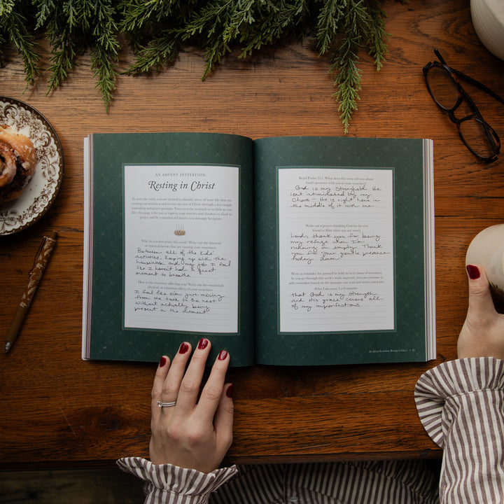 An open book with two printed pages titled 'Resting in Christ' laid on a wooden table, surrounded by holiday greenery, a pastry, eyeglasses, and a person's hands holding the book.