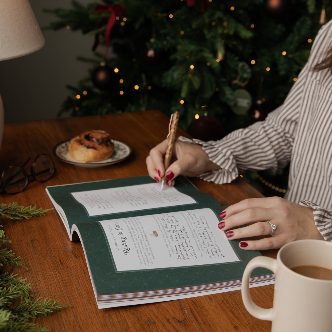 A person writing in an open green journal with a decorative wooden pen, set on a wooden table with a Christmas tree, a mug, and a pastry in the background.