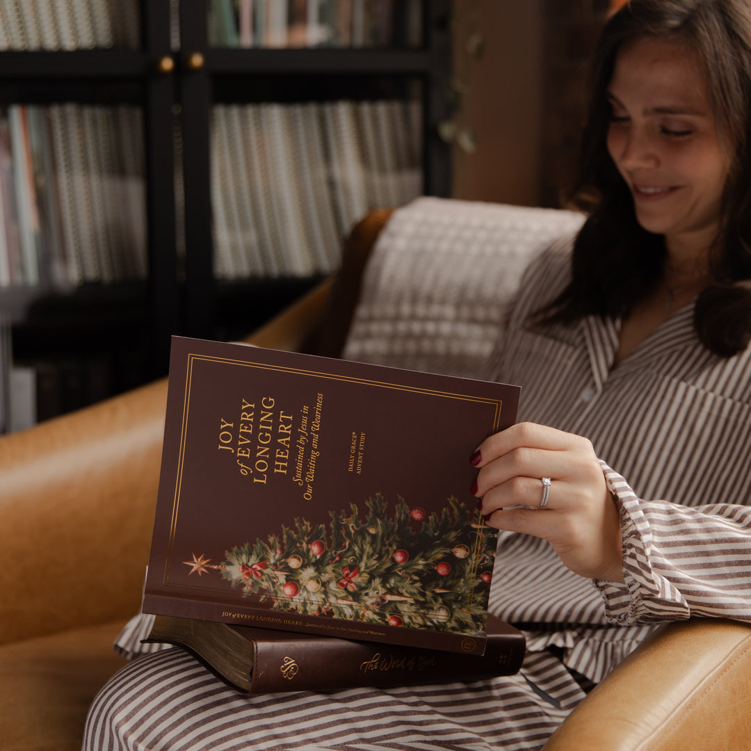A woman in a striped pajama shirt sits on a leather armchair holding a hardcover book titled 'Joy of Every Healing Hour' with a Christmas tree illustration on the cover.