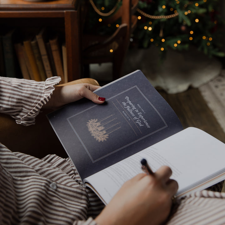 A person in a striped shirt holds an open book or journal in a cozy indoor setting with warm lights and a blurred Christmas tree in the background. The cover reads 'The Power of Prayer' with a gold illustration.
