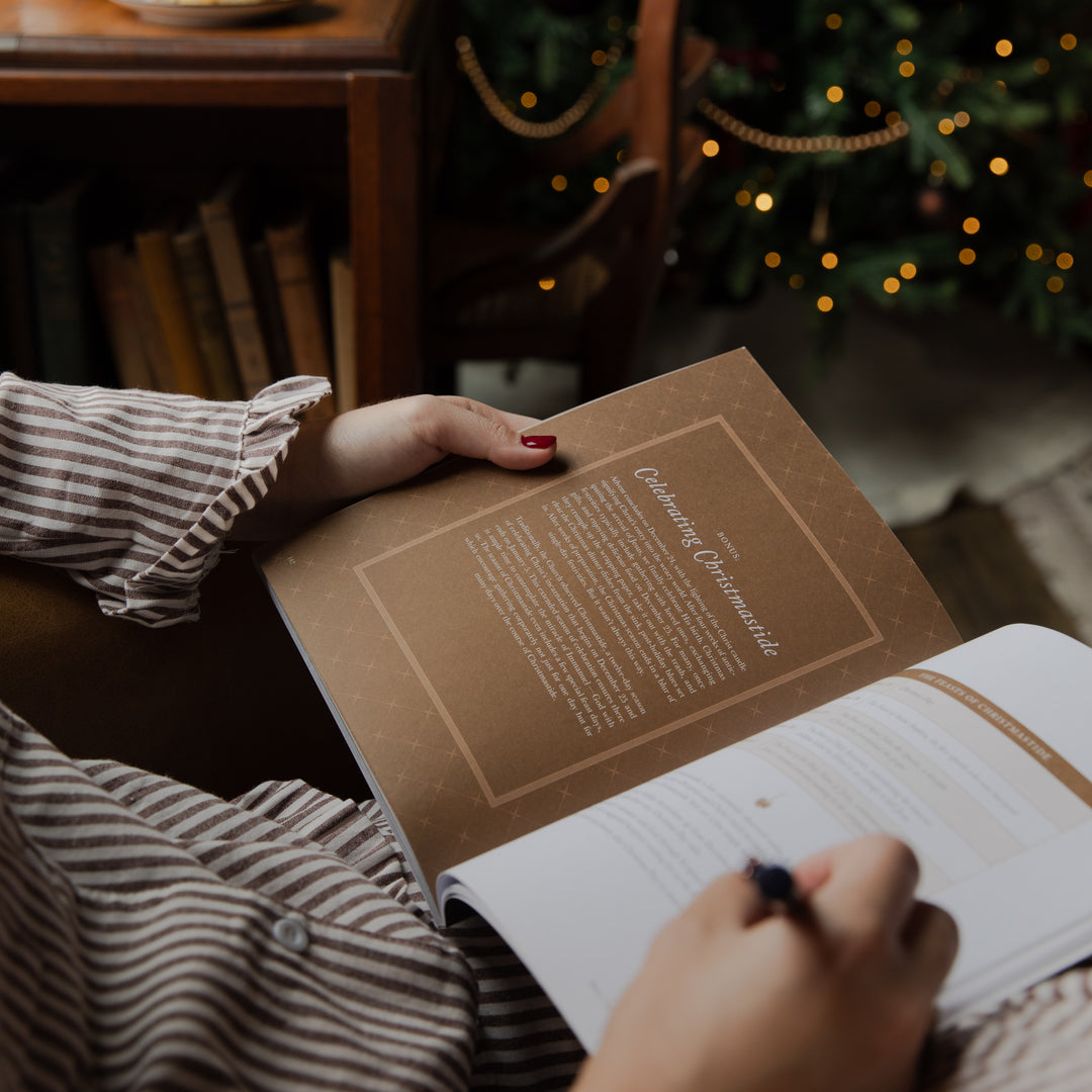 A person holding an open book or journal in a cozy indoor setting, with a blurred Christmas tree and warm lights in the background. The visible page has a brown cover and text, and the person is wearing a brown-and-white striped shirt.