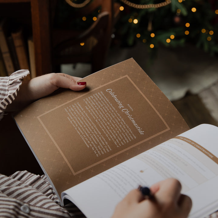 A person holding an open book with a brown cover page titled 'Celebrating Christmas' in a cozy indoor setting with warm holiday lights in the background.