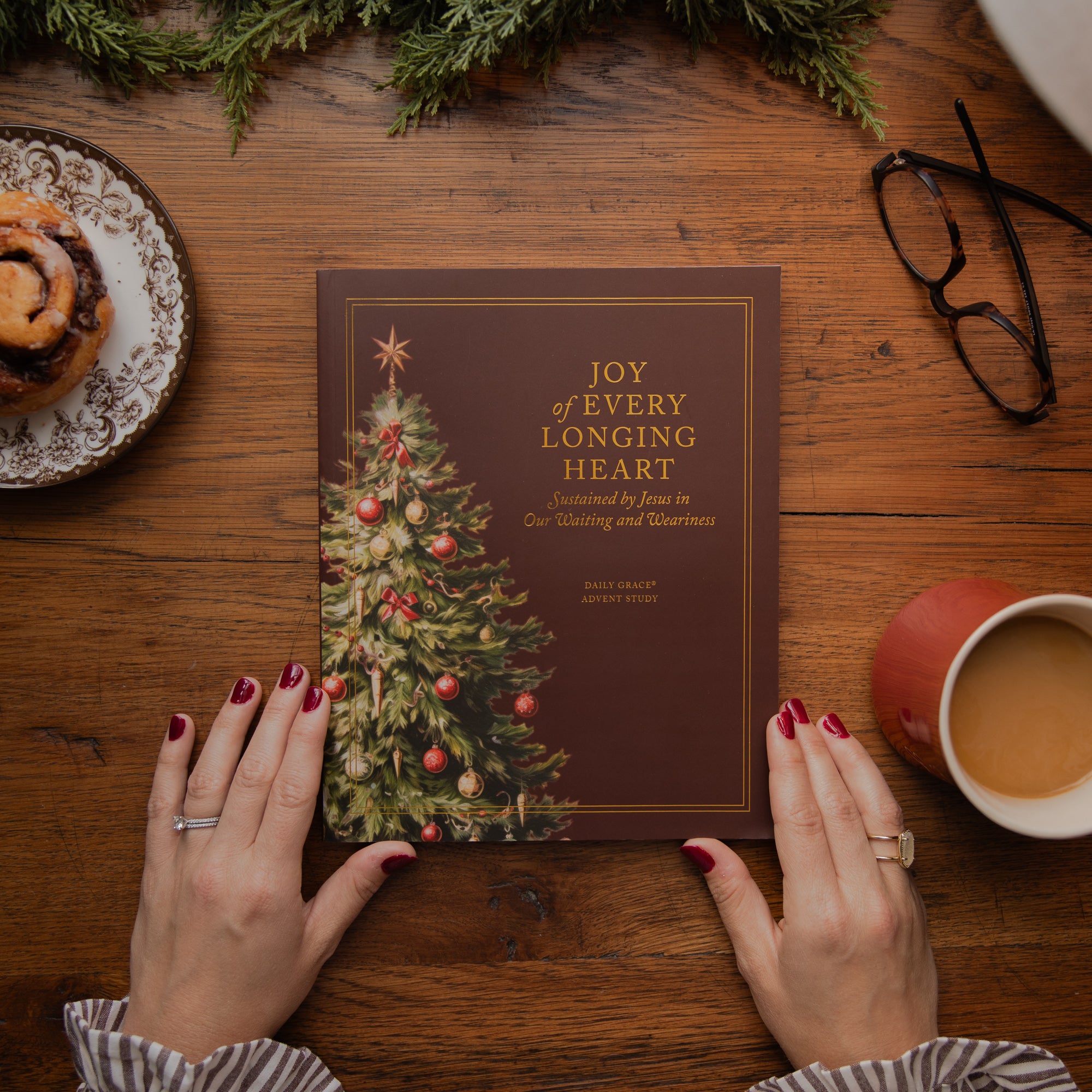 A person's hands with red nail polish hold a brown book titled 'Joy of Every Longing Heart' with a Christmas tree illustration. The scene is styled on a wooden table with a cinnamon roll, eyeglasses, a mug of coffee, and evergreen branches.