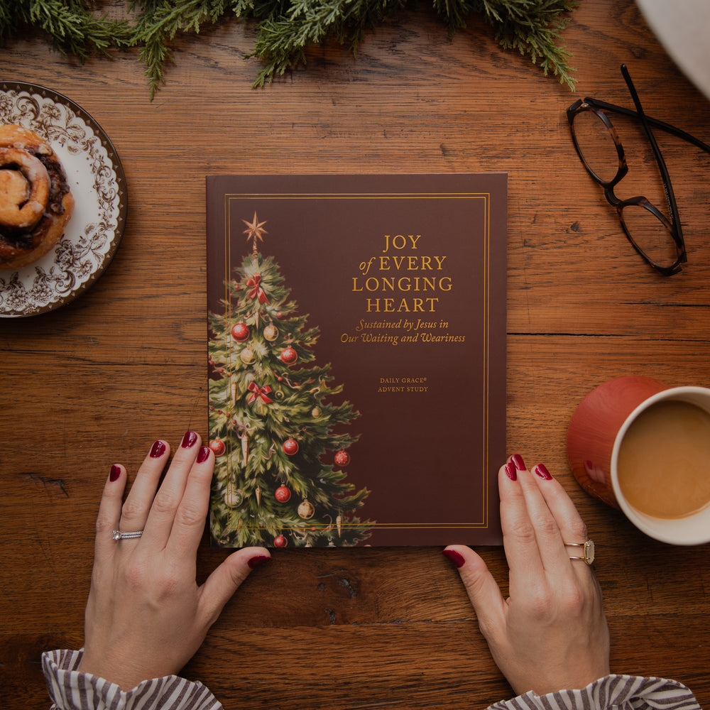 A person's hands with red nail polish hold a brown book titled 'Joy of Every Longing Heart' with a Christmas tree illustration. The scene is styled on a wooden table with a cinnamon roll, eyeglasses, a mug of coffee, and evergreen branches.