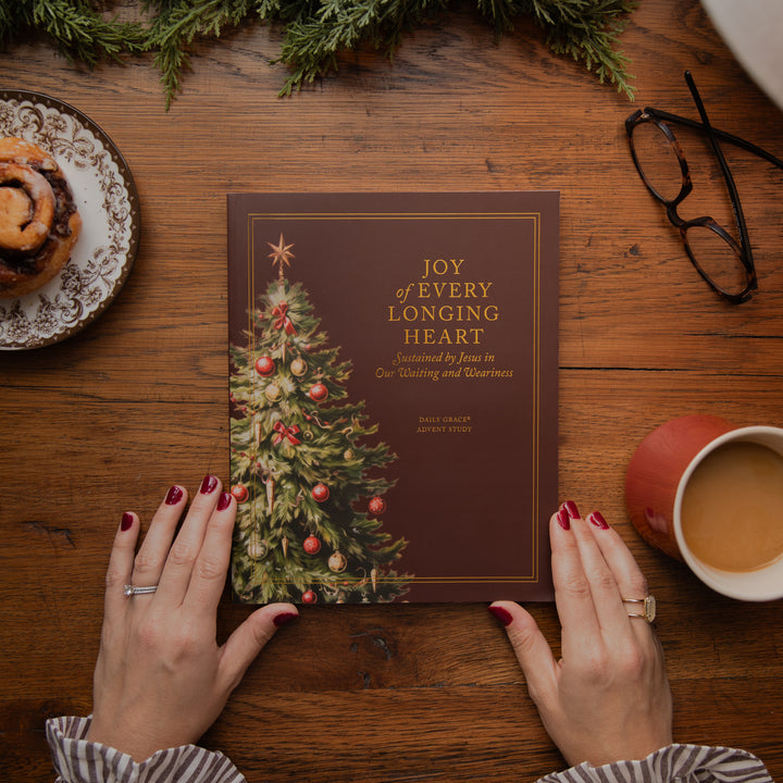 A person's hands with red nail polish hold a brown book titled 'Joy of Every Longing Heart' with a Christmas tree illustration. The scene is styled on a wooden table with a cinnamon roll, eyeglasses, a mug of coffee, and evergreen branches.