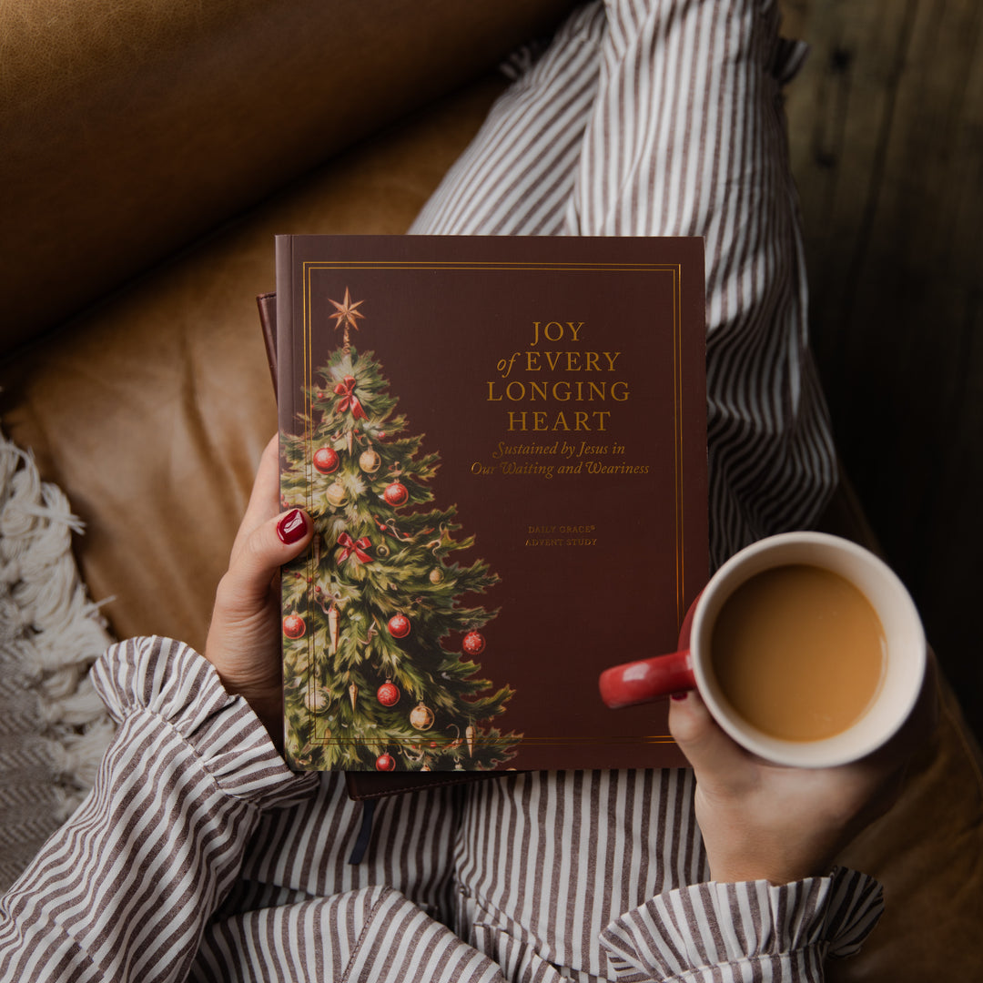 A person in a striped shirt holds a book titled "Joy of Every Longing Heart" with a Christmas tree illustration on the cover, while holding a mug of tea in a cozy indoor setting.
