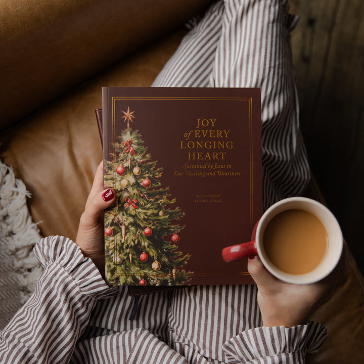 A person in a striped shirt holds a book titled "Joy of Every Longing Heart" with a Christmas tree illustration on the cover, while holding a mug of tea in a cozy indoor setting.
