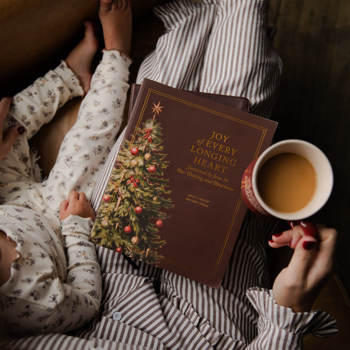 A cozy lifestyle scene showing a person in striped pajamas holding a red hardcover book titled 'Joy of Every Longing Heart' advent bible study with a Christmas tree illustration, while a baby in floral pajamas reaches toward the book and a hand holds a mug of tea.