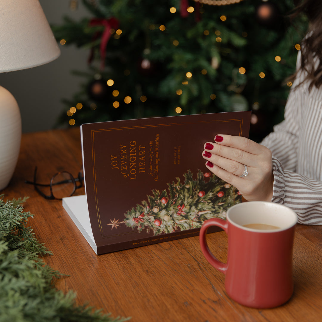 A person holds a brown book titled 'Joy of My Longing Heart' with a Christmas tree illustration on the cover, set on a wooden table next to a red mug, with a blurred Christmas tree in the background.