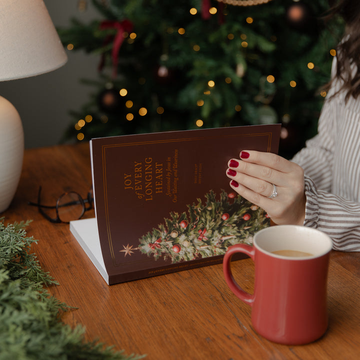 A person holds a brown book titled 'Joy of My Longing Heart' with a Christmas tree illustration on the cover, set on a wooden table next to a red mug, with a blurred Christmas tree in the background.